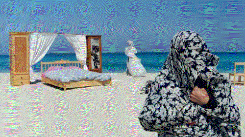 A woman with a shawl wrapped over her head stands on a beach, with furniture behind her, in this still from The Day I Became a Woman.
